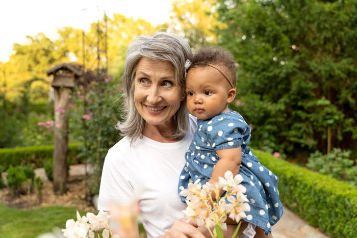 Smiling grandmother holding a baby in a polka-dot dress, surrounded by greenery. Grandkid's allergies are a concern.
