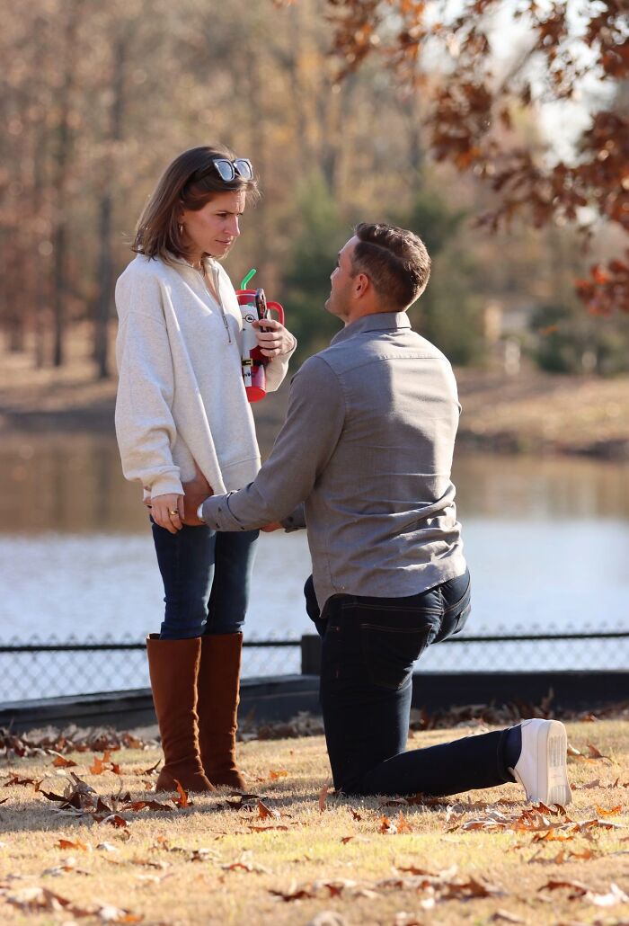 Man proposing on one knee to a surprised woman by a lake, a romantic engagement moment. Captures love and chaos.