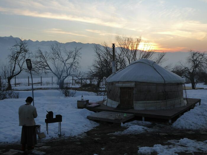 Winter scene with a yurt on a raised deck, a person cooking outdoors, and mountains at sunset. Perfect for hilarious engagement stories.