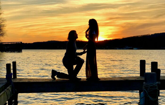 Silhouette of a man proposing to a woman on a dock at sunset, perfect for engagement stories and romance.
