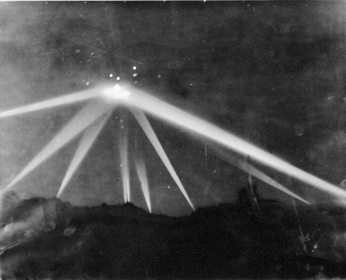 Black and white photo showing bright beams of light crossing night sky over dark landscape