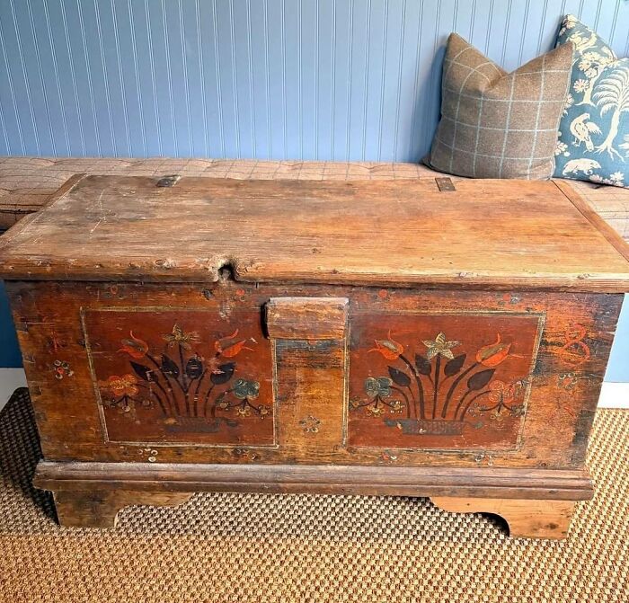 An historic piece from early America: a rustic wooden chest with painted floral designs, sitting on a woven rug.
