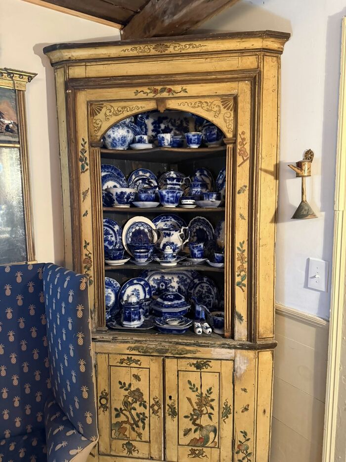 Corner cabinet filled with historic pieces from Early America, featuring blue and white dishes and decorative paintings.