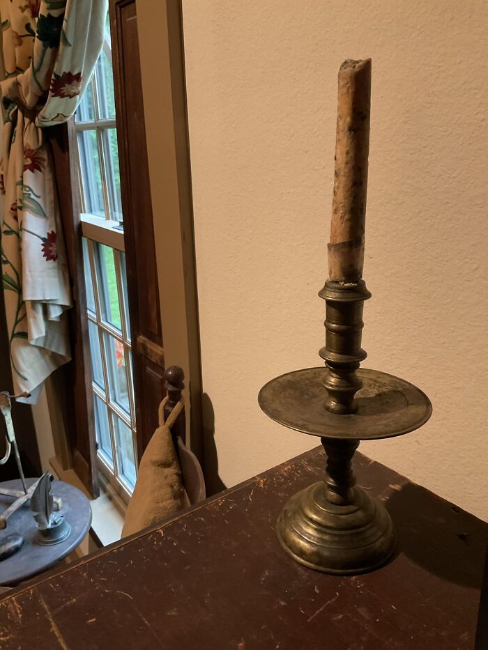A vintage brass candlestick with a partially burnt candle on a wooden table, near a window with floral curtains. Early America antique.
