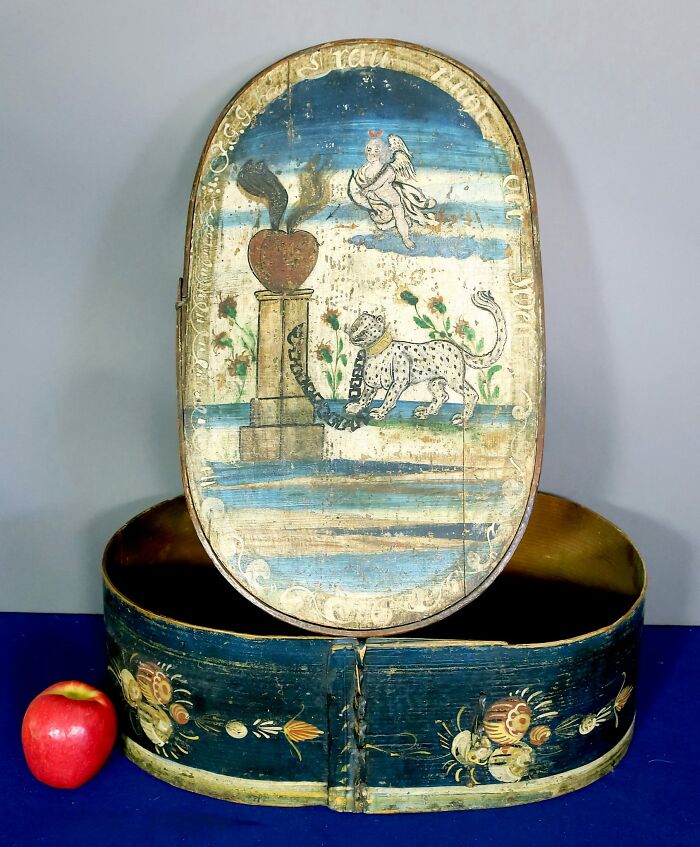 A painted oval wooden box, open to reveal its colorful lid. An historic piece from early America, with a leopard and cherub.