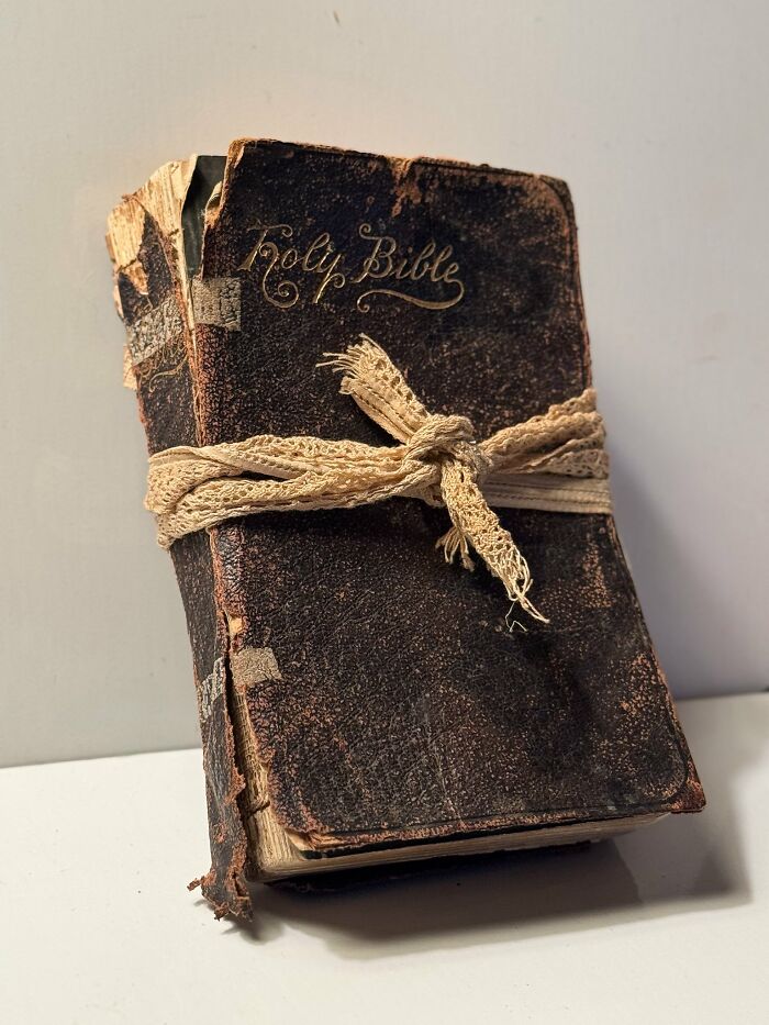 An old, worn Holy Bible with a dark brown cover and faded gold lettering, tied with a cream lace ribbon. Historic piece.