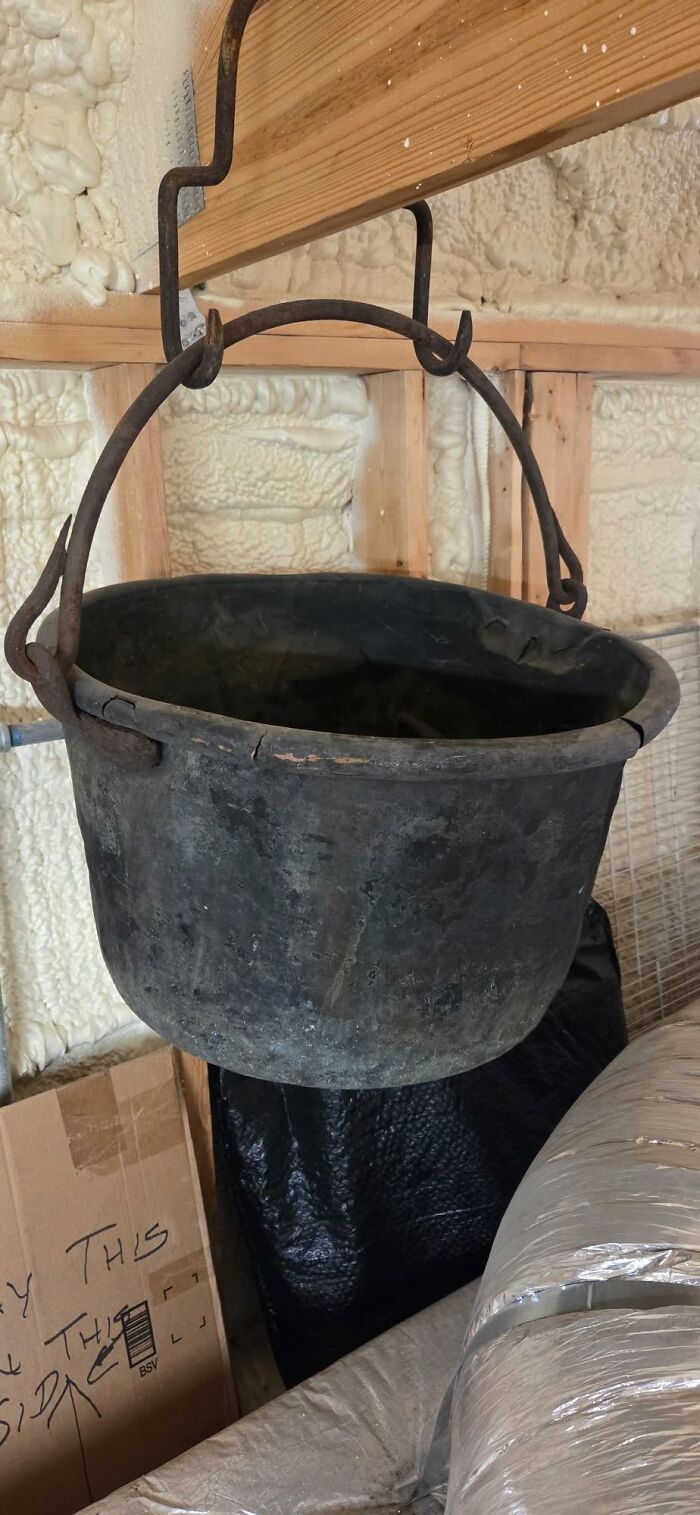 A tarnished, black cauldron with a rustic metal handle hangs from a wooden beam. A historic piece from early America.