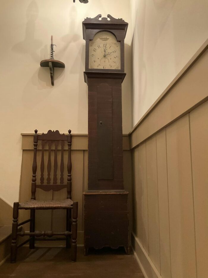 An antique wooden grandfather clock stands tall next to a colonial-style chair, showcasing historic pieces from early America.