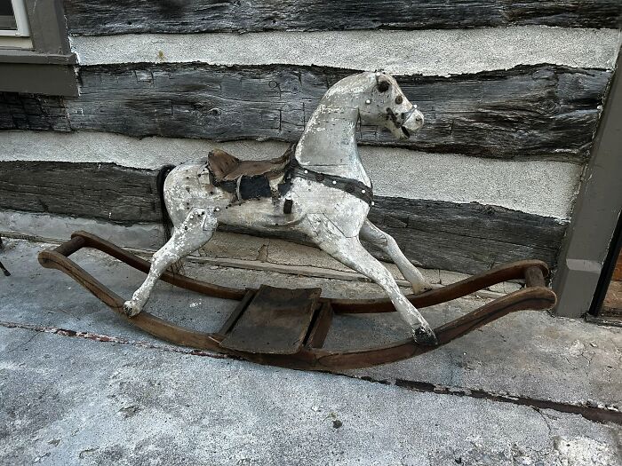 An old, white, weathered rocking horse, an historic piece from Early America, against a rustic log cabin wall.