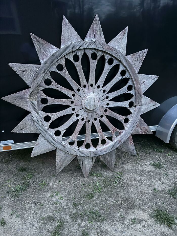 An historic piece from early America, this large, weathered wooden wheel with decorative cutouts and pointed spokes, rests on grass.