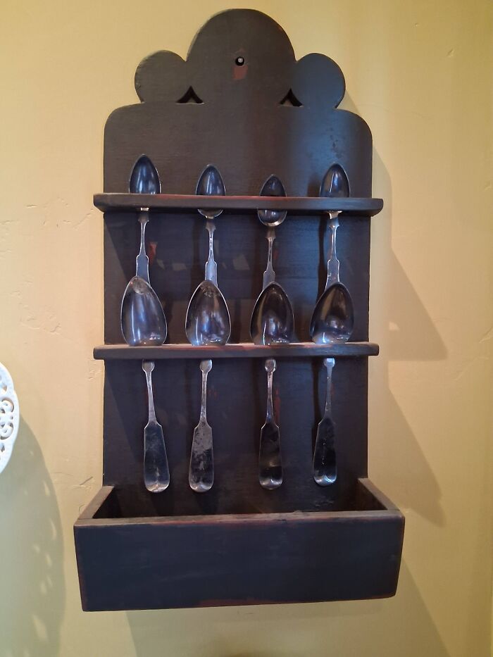 A wooden spoon rack with eight early America collectible spoons and ladles, two rows of four on a yellow wall.