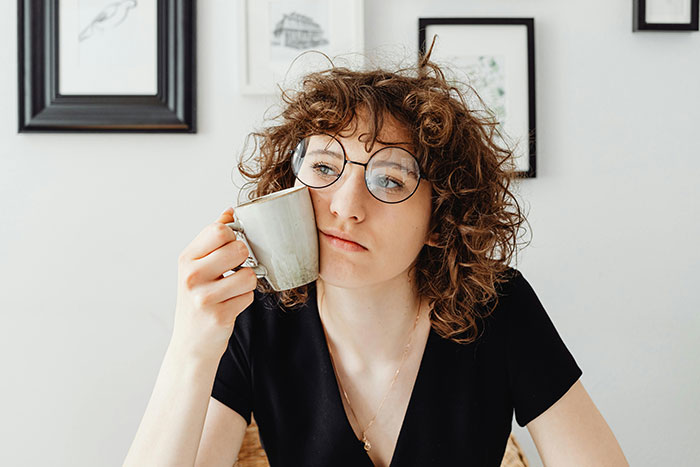 A young woman with curly hair and round glasses, looking pensive while holding a coffee cup to her cheek. No kids.