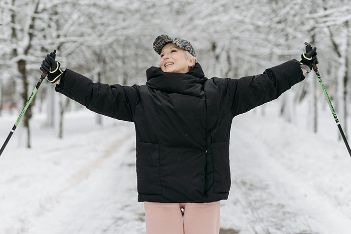 Happy older person, dressed in a winter coat and cap, enjoying Nordic walking in a snowy park. Not to have kids.