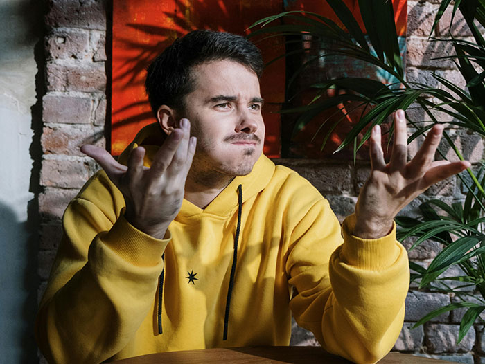 Man in yellow hoodie expressing disbelief indoors with plants and brick wall Man in yellow hoodie expressing disbelief indoors with plants and brick wall