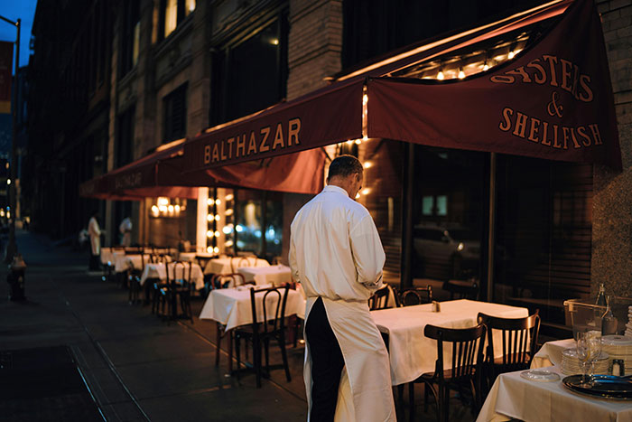Man standing outside Balthazar restaurant at night with empty outdoor tables Man standing outside Balthazar restaurant at night with empty outdoor tables