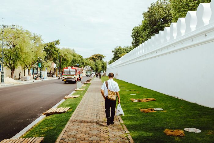 A man walking on a sidewalk next to a long white wall and green grass, with a red bus on the street. Science professionals.