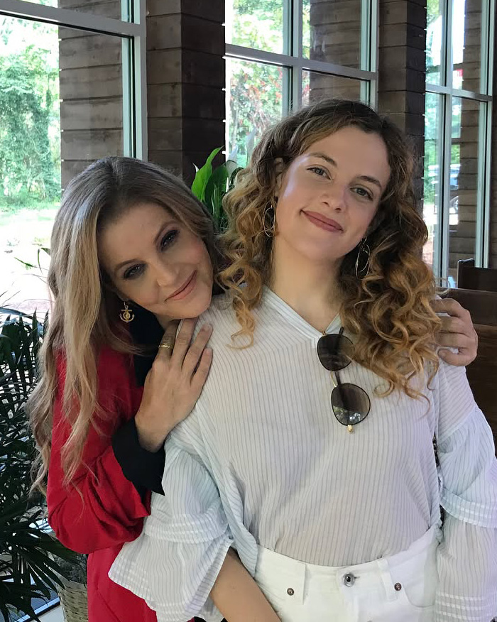 Priscilla Presley (left) in a red top, with her arm around a young woman (right) with curly hair. Fans slam Priscilla Presley.