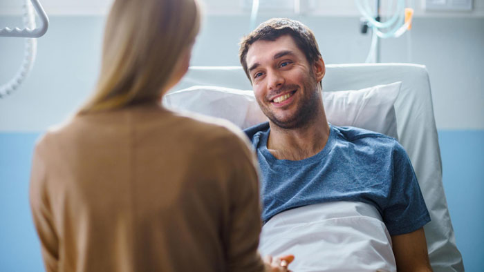 A SAHM visiting her fianc&eacute; in a hospital bed, where he is smiling. They are discussing his debts.