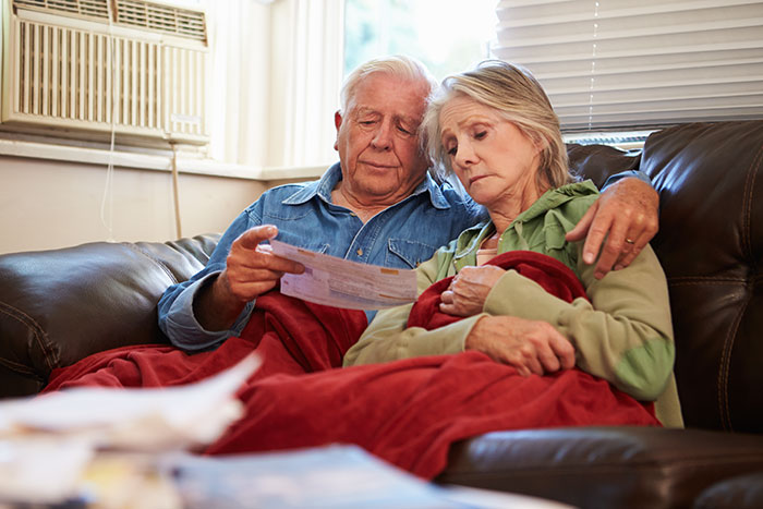 An older couple, under a red blanket on a couch, looking at papers. The man supports the woman, reflecting manipulative parents.