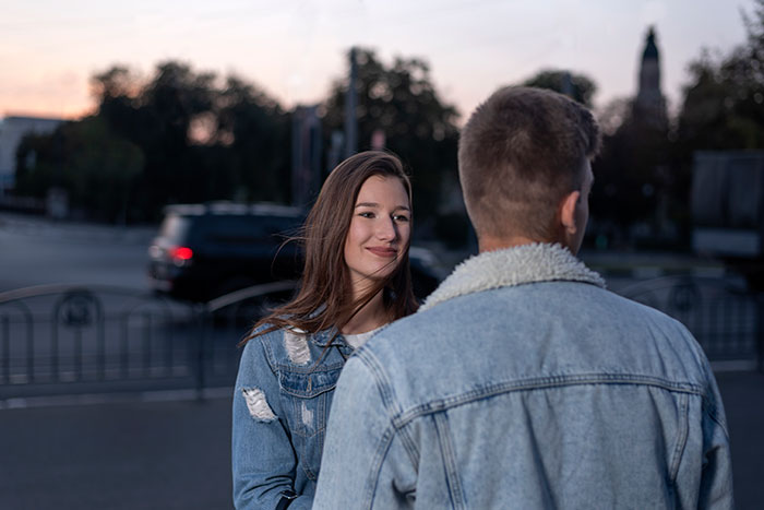 A young woman with a slight smile looks at a man's back, a first date interaction in a city at dusk.