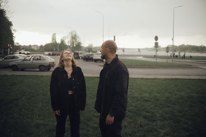 A young couple, the woman laughing, stand in a park near a street. Reflecting cringe-worthy first date stories.