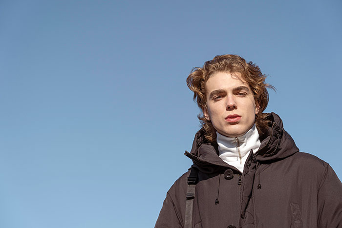 A young man with wavy hair and a black winter coat against a blue sky, reflecting some cringe-worthy date stories.