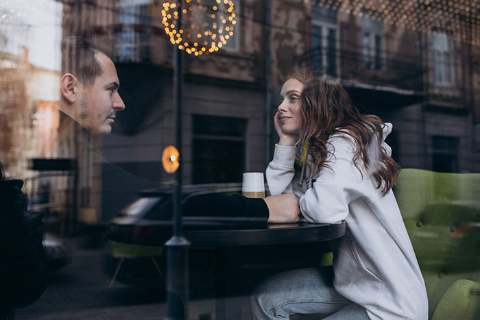 A man and woman on a first date, seen through a window reflecting the city. She looks thoughtful, possibly a cringe-worthy or scary first date story.