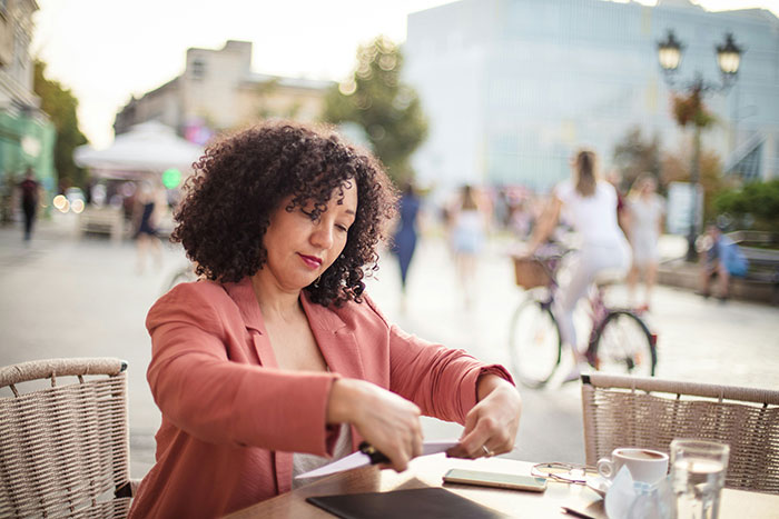 A woman with curly hair sits at an outdoor cafe, focused on paperwork, with a street scene blurring behind her. Scary first date stories.
