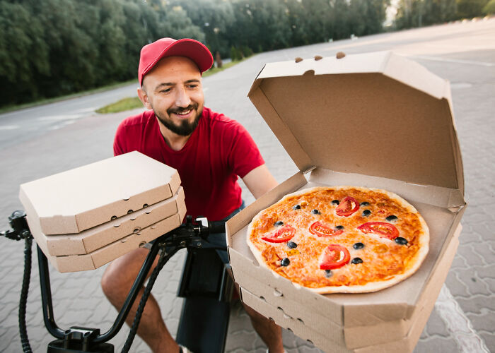 Pizza delivery driver holding pizza boxes and showing a pizza on bike outdoors