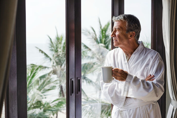 Older man in robe holding coffee cup looking out window