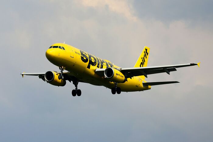 A Spirit Airlines plane in flight, its bright yellow fuselage and black "Spirit" logo visible against a cloudy sky.