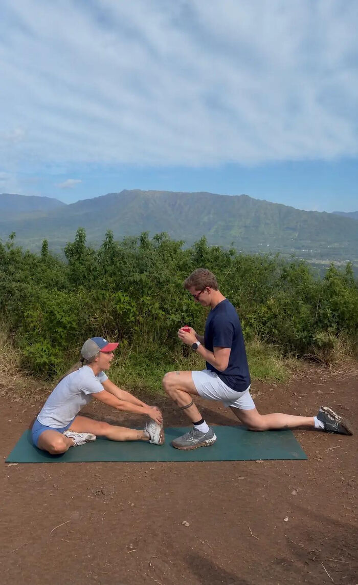A woman stretching on a mat and a man kneeling, holding a red box, in a proposal pic with mountains in background.