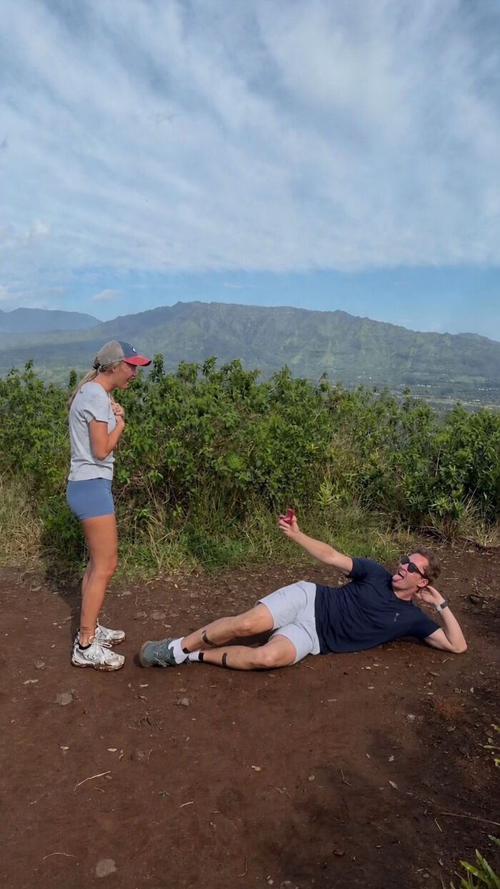 A woman looking surprised while a man lies on the ground, holding a proposal pic with a mountain background.
