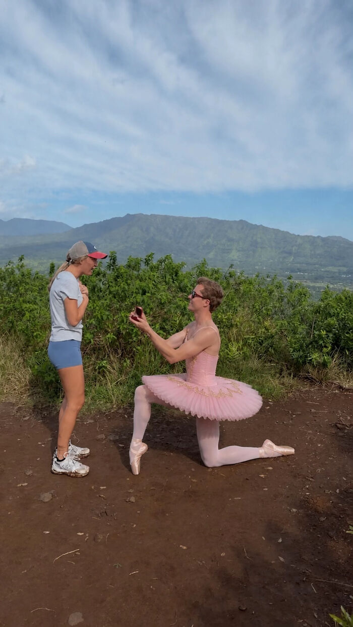 A man in a pink tutu and pointe shoes proposes to a woman on a hill overlooking mountains. Hilarious edits of this proposal pic are popular online.