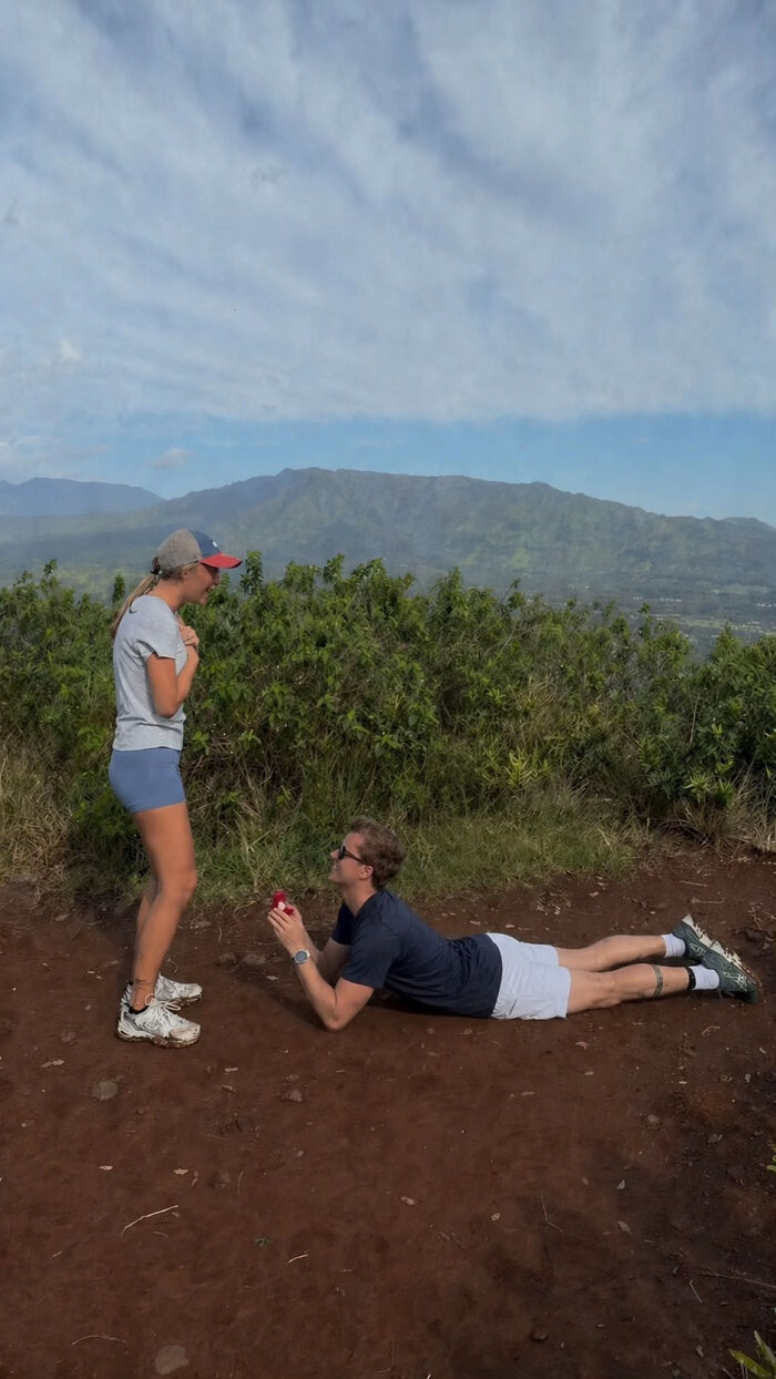 Woman reacting to a man's proposal pic, lying down with a ring box, on a hiking trail overlooking mountains.