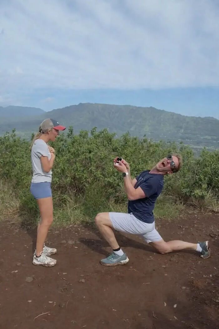 A man in sunglasses and a blue shirt kneels to propose to a woman on a dirt path with green mountains in the background. His proposal pic sparked hilarious edits.