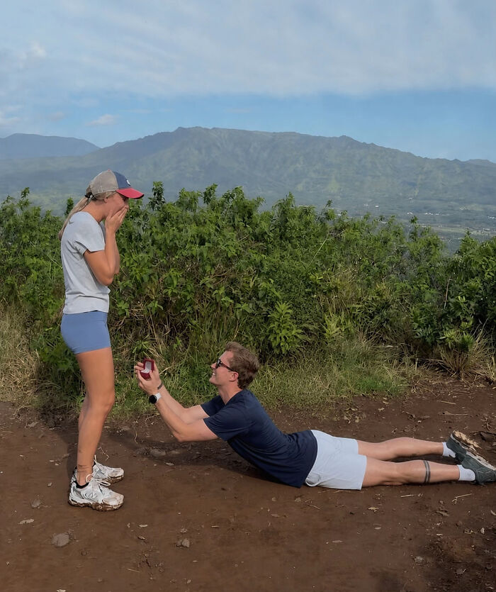 Man proposing to woman in a memorable proposal pic, lying on the ground, holding a ring box, with mountains in background.