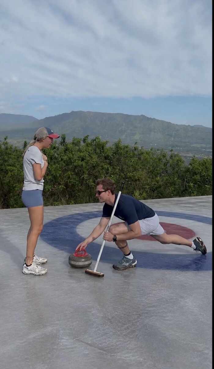 A man makes a curling proposal, kneeling with a curling stone. A woman watches, surprised, outdoors on a sunny day. This proposal pic is hilarious.