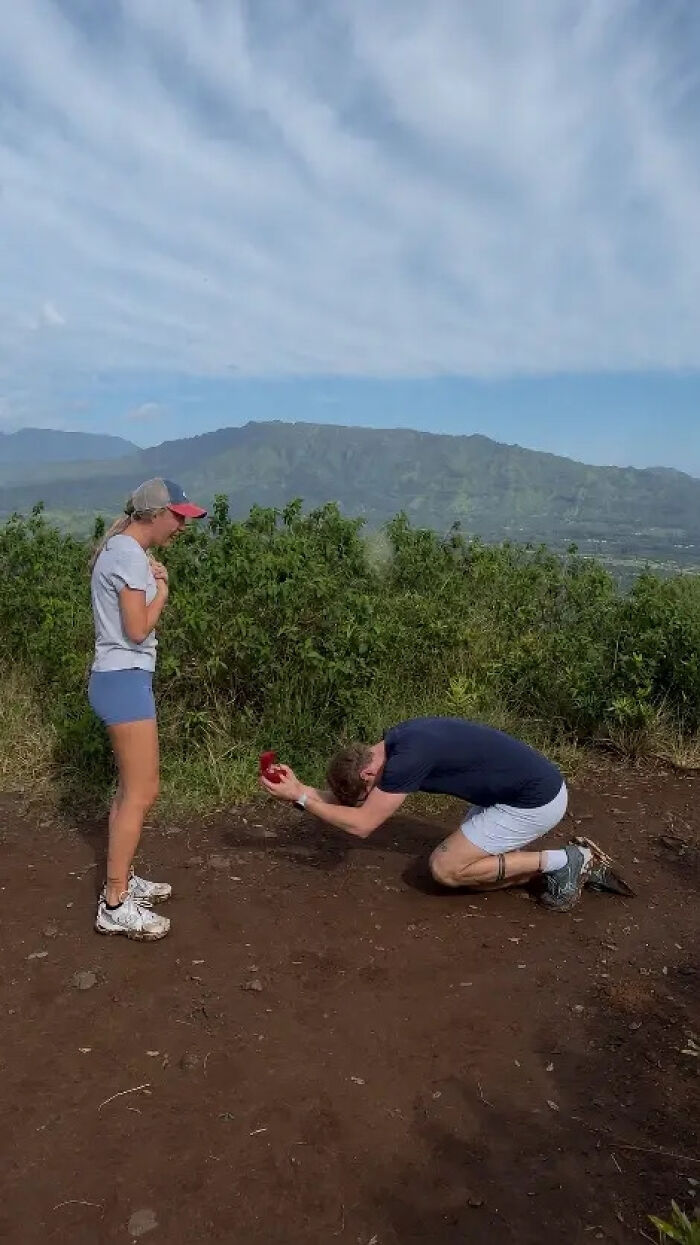 Man kneels to propose to a woman on a scenic mountain path. A memorable proposal pic that inspired hilarious edits.