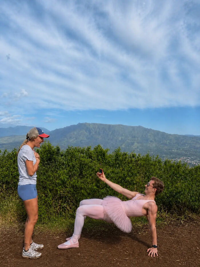 Man in a pink tutu and tights proposes to a woman who looks surprised. A hilarious proposal pic with mountains in background.