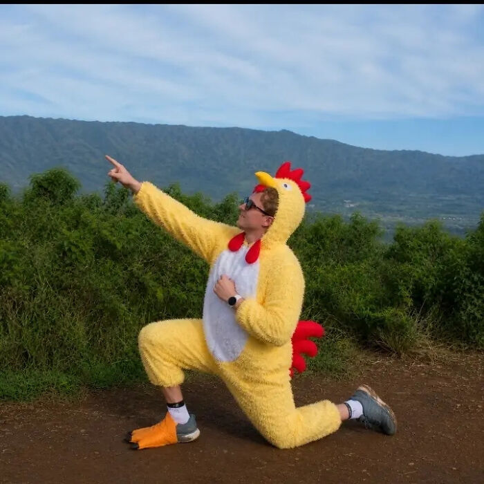 A person in a chicken suit, kneeling and pointing, with a mountain landscape behind them, a funny proposal pic edit.