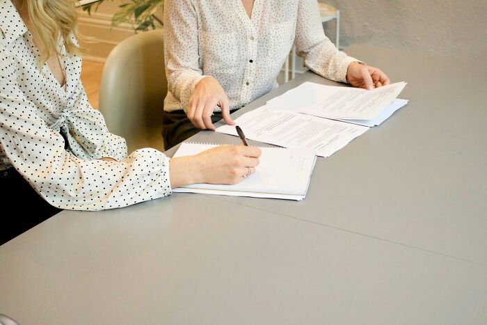 Two women in polka dot blouses at a table, one taking notes, discussing rich people's silent luxuries.