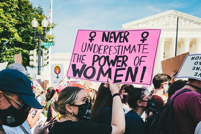 A woman holds a pink sign that says "Never Underestimate the Power of Women!" among a crowd of protestors. This image relates to the manosphere content discussion.