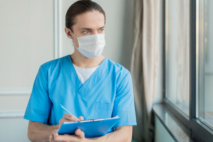 A medical professional in blue scrubs and a mask, holding a clipboard and pen, looking away thoughtfully. Wild encounters can sometimes challenge our intelligence.