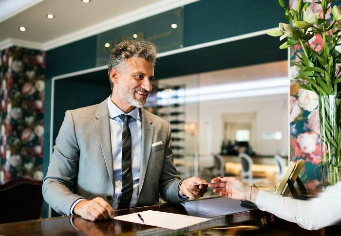 A smiling businessman in a grey suit and tie at a hotel reception, handing over a card. Catching BF cheating stories.