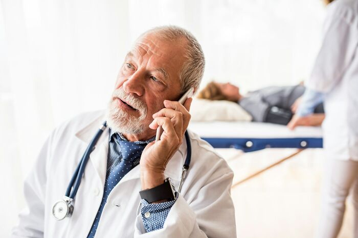 A senior male doctor in a white coat and stethoscope talks on a phone, with a patient in the background. Epic comebacks.