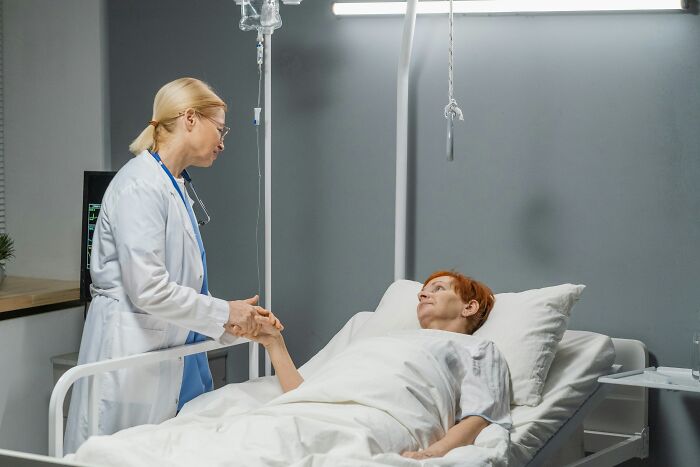 A doctor in a white coat holds the hand of a red-haired patient lying in a hospital bed, discussing routine cancer screening.