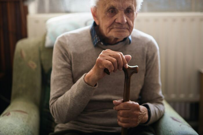 Elderly man in a sweater sitting, holding a walking stick. Reflecting on disturbing d***h stories.