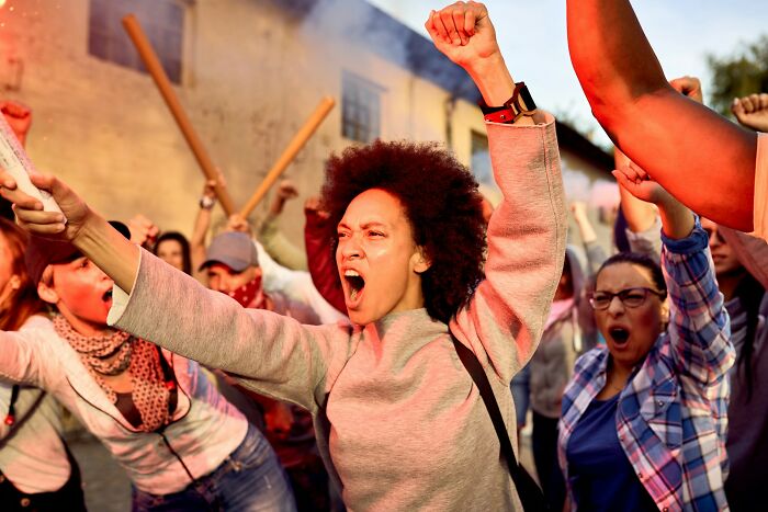 A diverse crowd of angry people, including a woman with curly hair yelling, at a protest. Highlights men suffering and manosphere content.