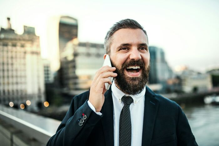 Um homem barbudo de terno ri enquanto fala ao telefone, parecendo alguém contando uma história sobre ter flagrado um namorado traindo.